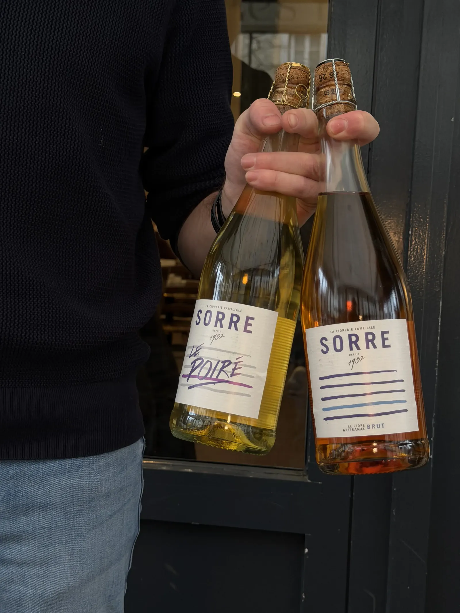 Bouteilles de poiré et de cidre brut fermier Sorre tenues devant la façade de la crêperie