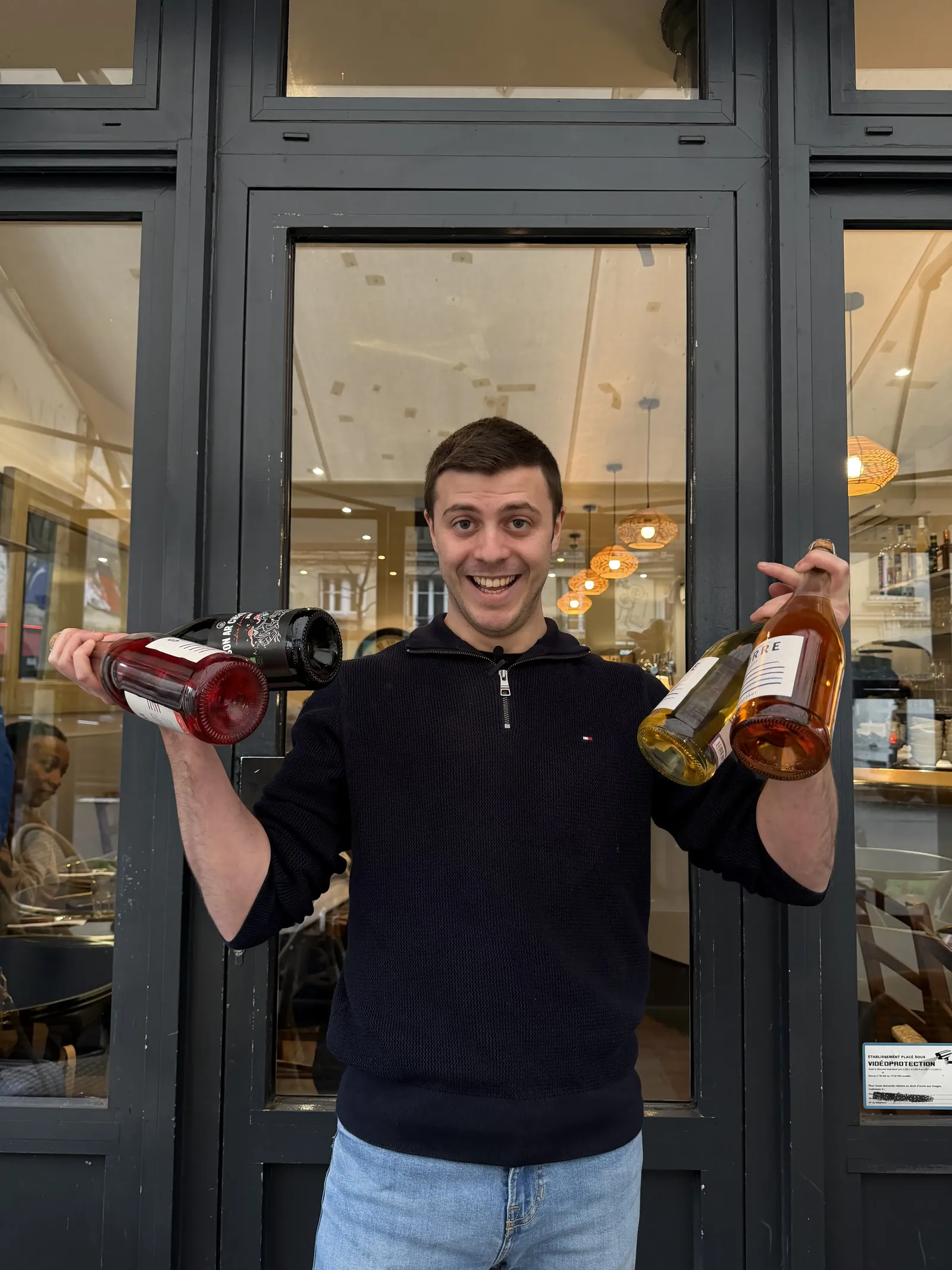 Membre de l’équipe souriant devant la façade des Embruns, deux bouteilles à la main